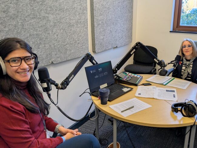 2 people sitting at a table with large microphones looking at the camera
